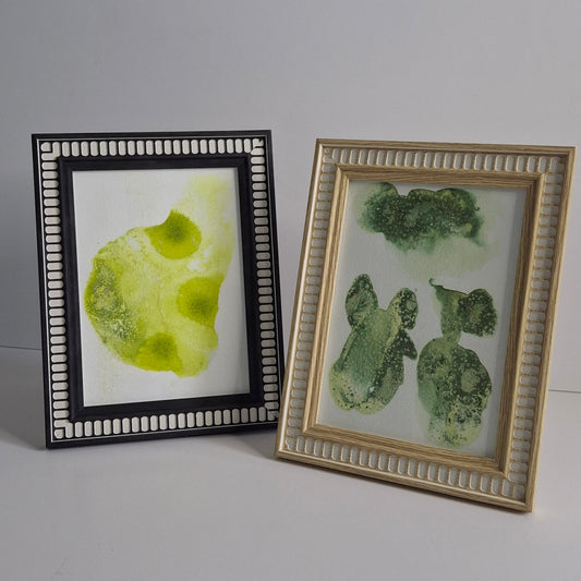 Two small green abstract artworks framed in patterned timber frames. Left piece features soft lime shapes; right piece has textured green botanical forms.