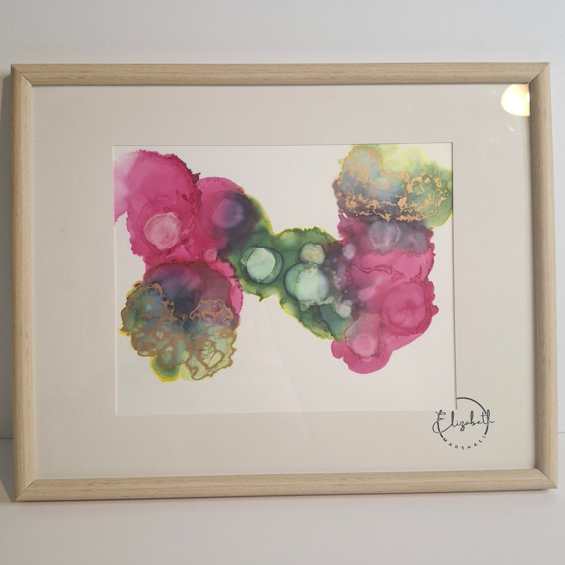 Framed abstract alcohol ink artwork titled Connected We Bloom by Elizabeth Marshall, featuring overlapping circular forms in rich pink, green, and teal tones with gold detailing. Displayed in a pale wooden frame against a white wall.