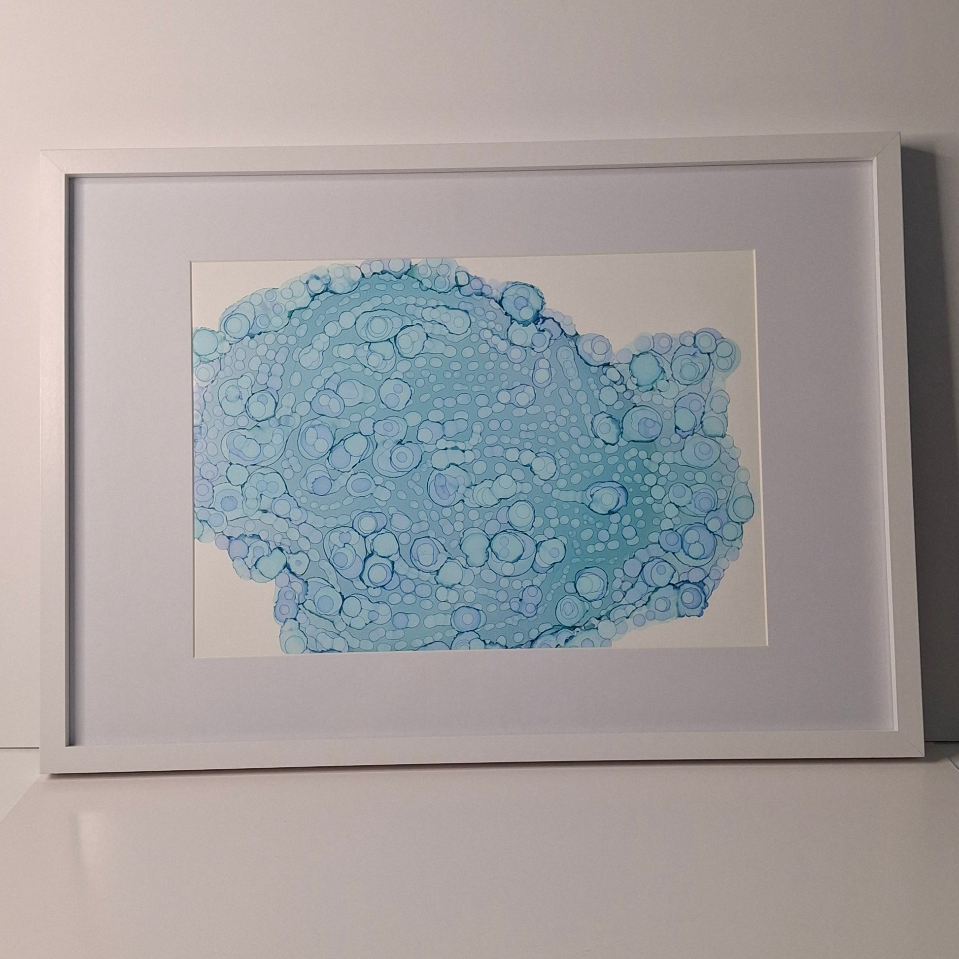 Framed blue alcohol ink artwork with overlapping bubbles and oceanic textures titled The Sea's Soft Embrace displayed in a coastal white kitchen